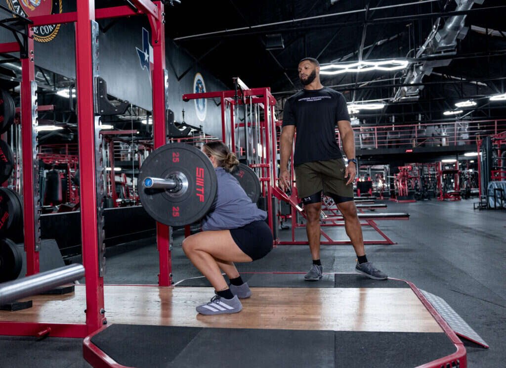 Woman squatting a loaded barbell as a personal trainer watches at Gryffon Performance in Dallas